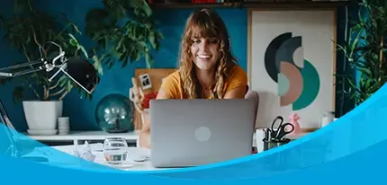 A smiling woman works on a laptop at a white desk while a black dog rests on a rug nearby.